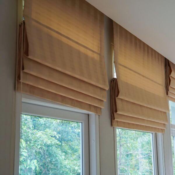 Roman blinds in a warm tan fabric, partially raised to show soft stacked folds across tall windows, creating filtered natural light, installed by Blinds and Shutter Guys Newcastl